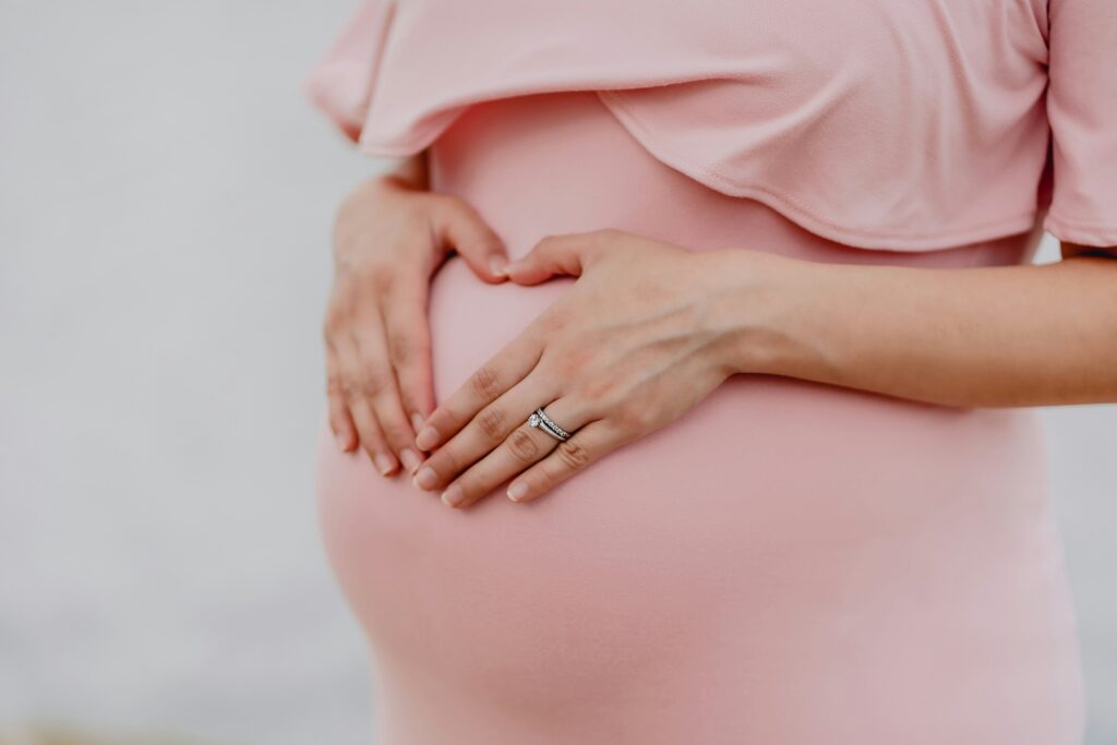 Endometriose: Eine schwangere Frau im rosa Kleid legt die Hände schützend auf Ihren Bauch.